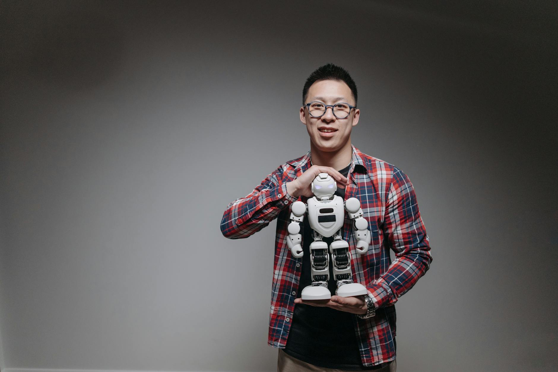 Asian man with eyeglasses holding a toy robot in a studio with a gray background.