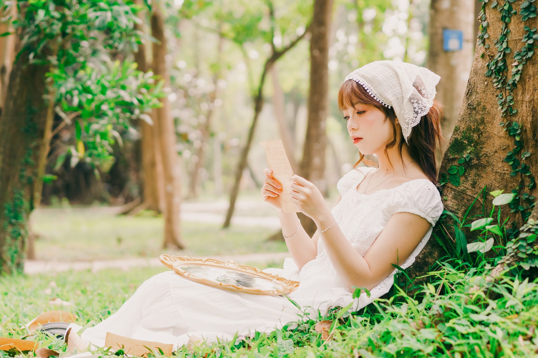A young woman in vintage attire reads a letter while sitting against a tree in a serene Hanoi park.