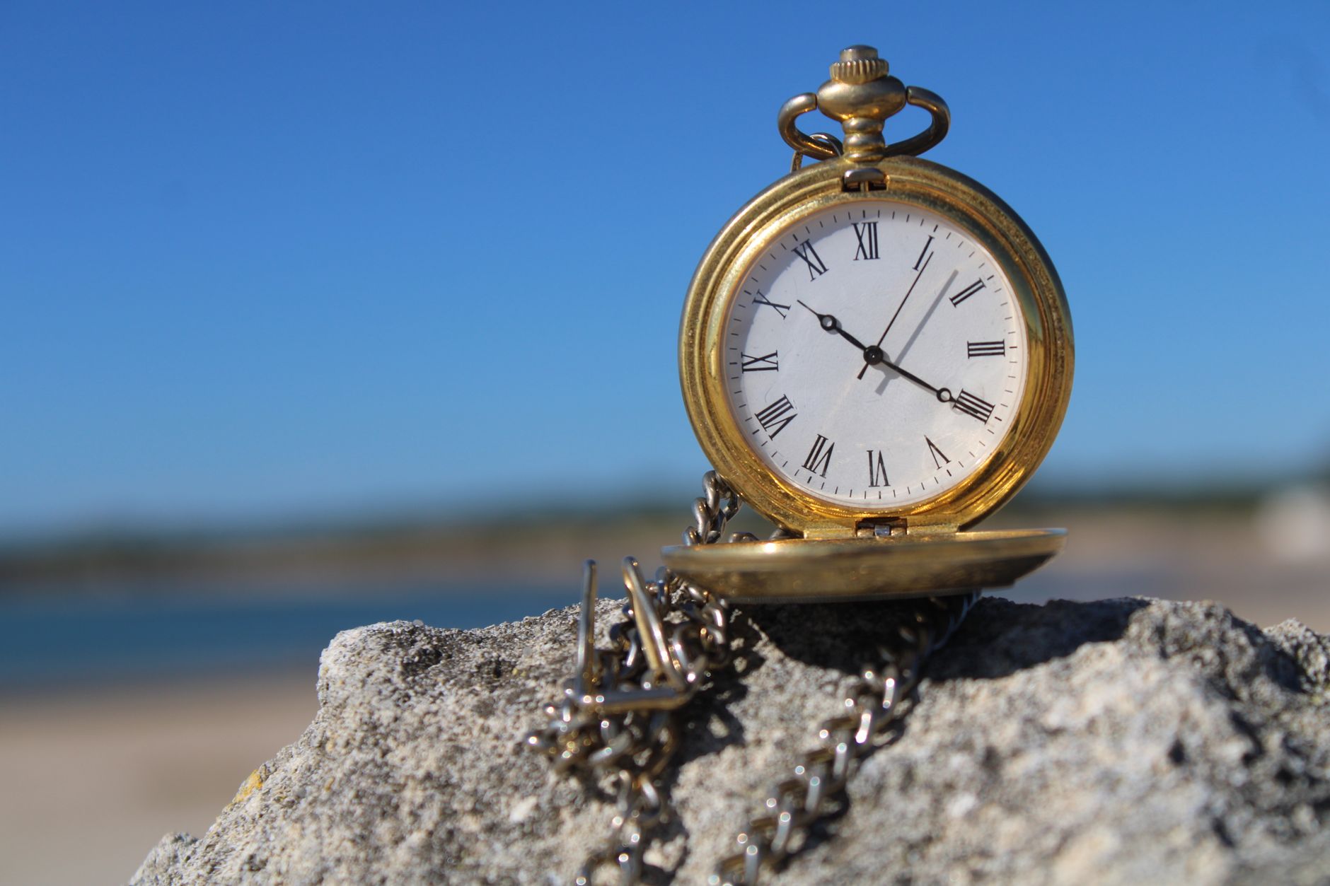 A classic gold pocket watch with Roman numerals sitting on a coastal rock under a clear blue sky.