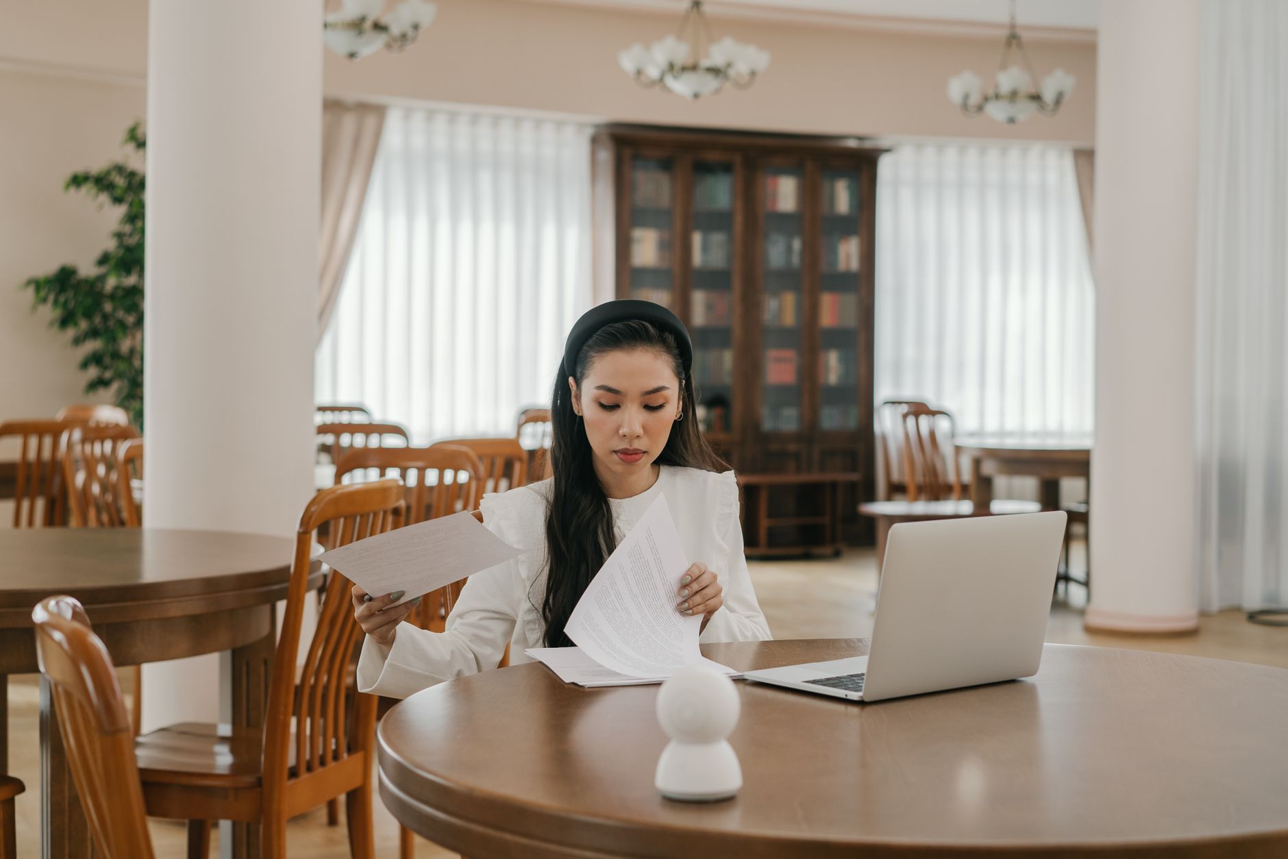Asian woman using laptop for online learning in a modern library setting, surrounded by books and wooden furniture.