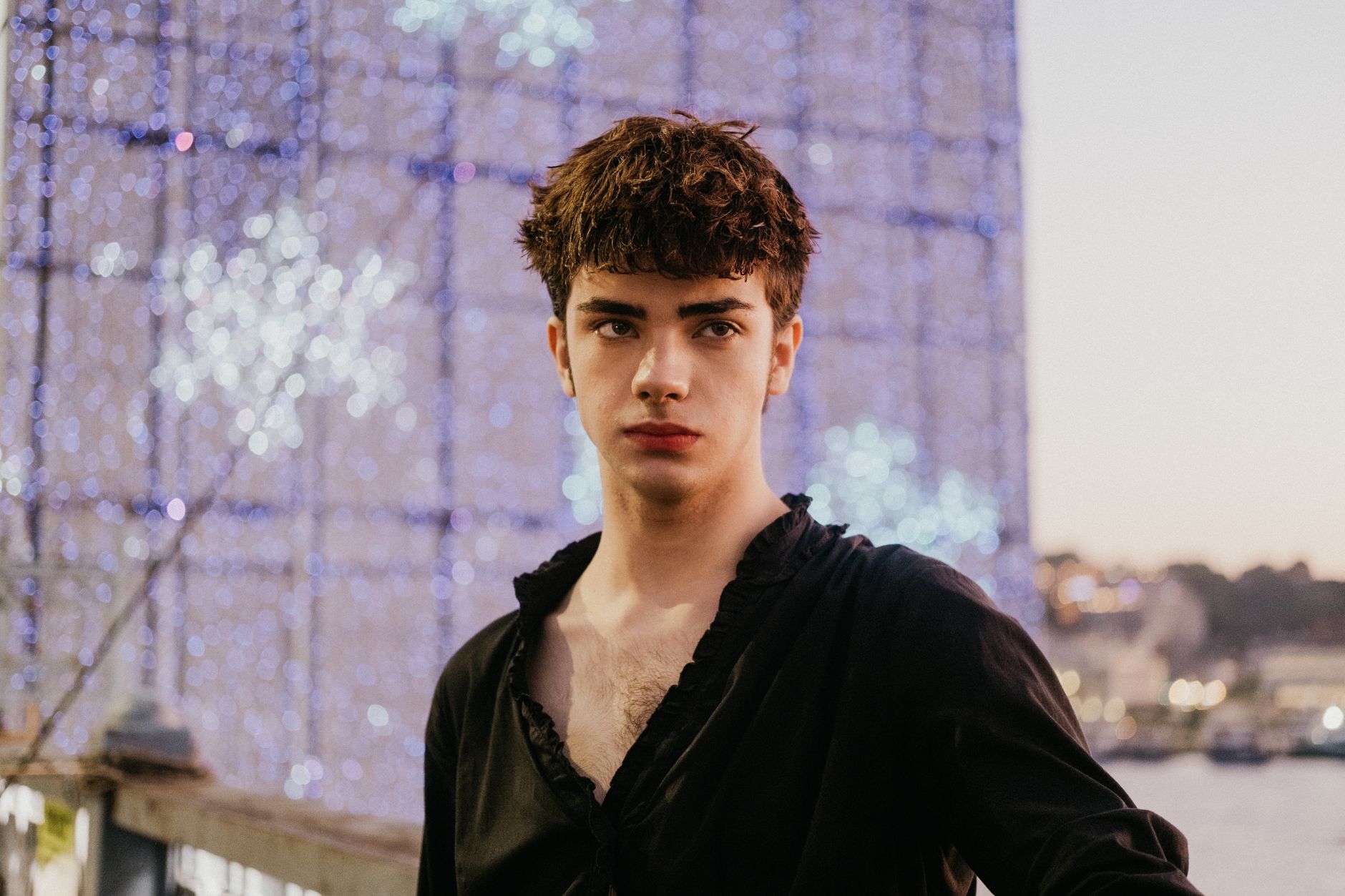 A young man in a black shirt posing outdoors in İstanbul with vibrant night lights.